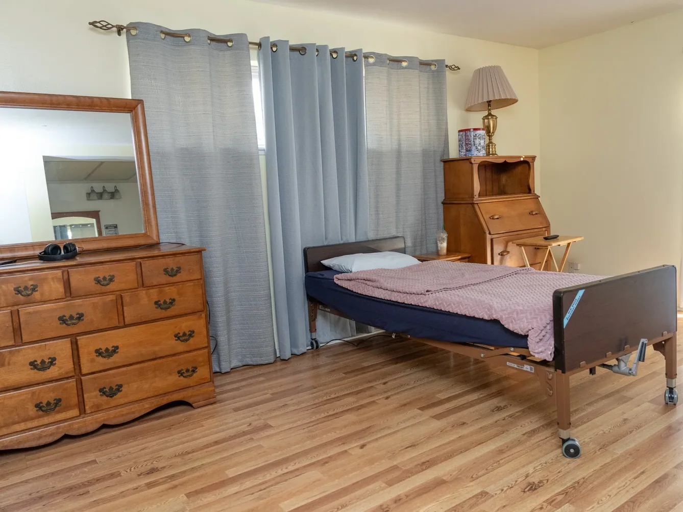 Spacious accessible bedroom featuring hospital bed with mauve bedding, wooden dresser with mirror, and traditional furnishings in warm neutral tones.